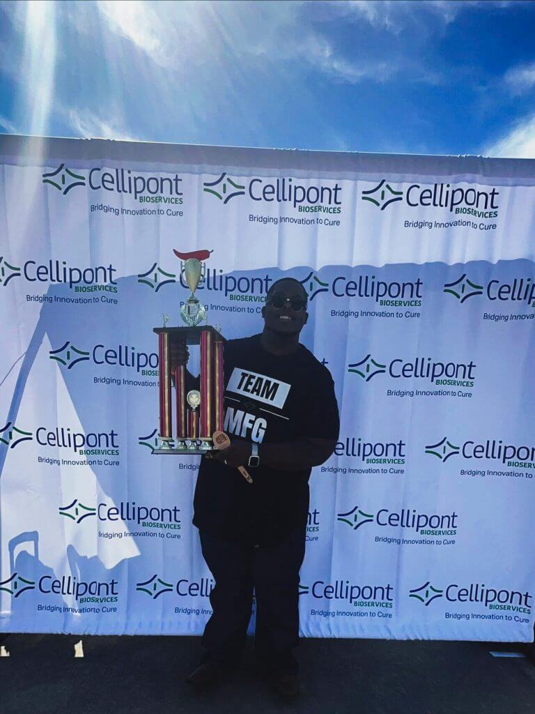 Cellipont Bioservices employee in a “TEAM MFG” shirt smiling and holding a large trophy and wooden spoon in front of a branded backdrop.