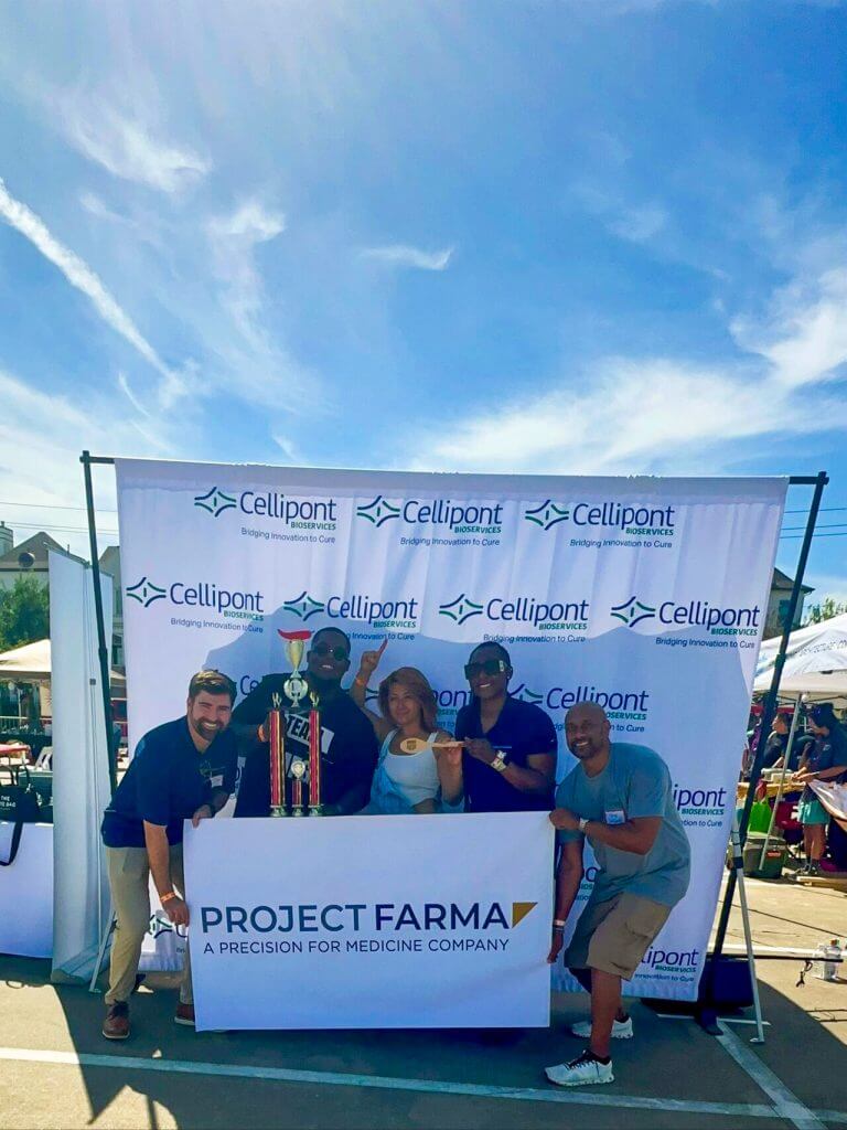 A group of people posing with a trophy and wooden spoon in front of a Cellipont Bioservices and Project Farma banner at an outdoor event.